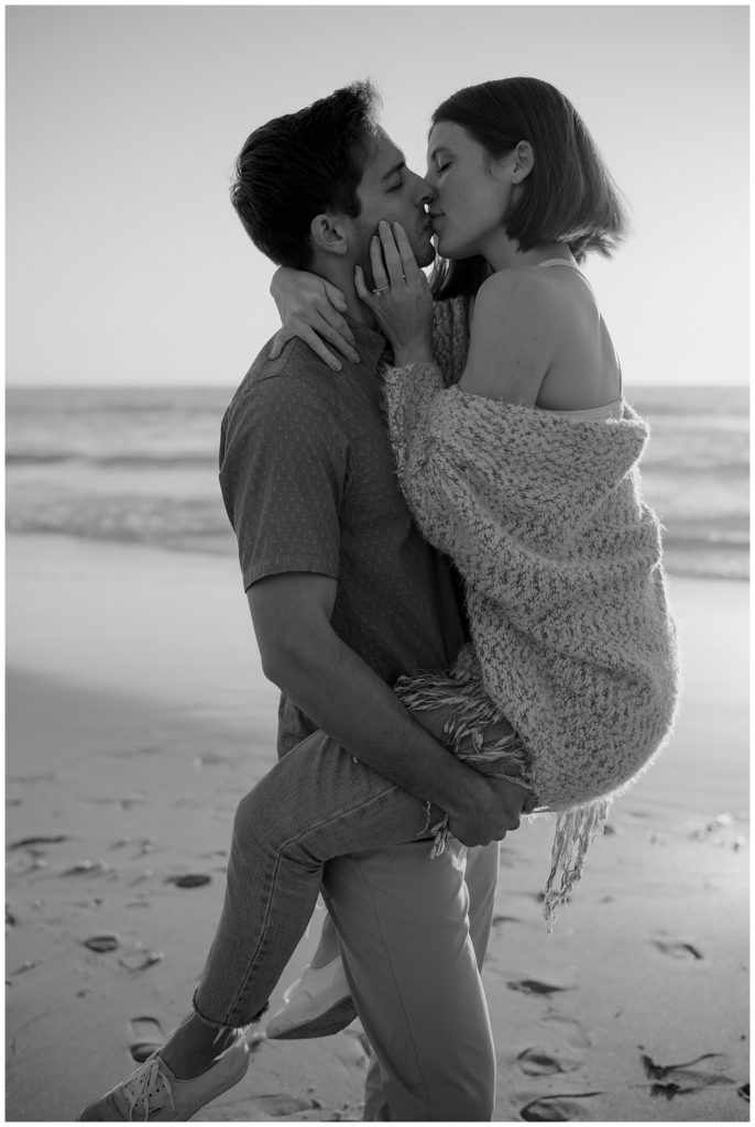 man holding woman in beach seaside engagement session location - black and white couples photo